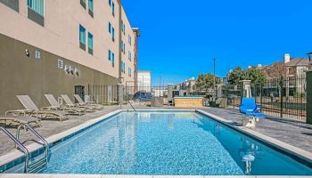 An outdoor swimming pool surrounded by lounge chairs, a modern building, and clear blue skies in the background.