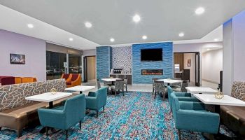 A modern lounge area features bright blue and neutral seating, a patterned carpet, multiple tables, a wall-mounted TV, and a refreshment station.