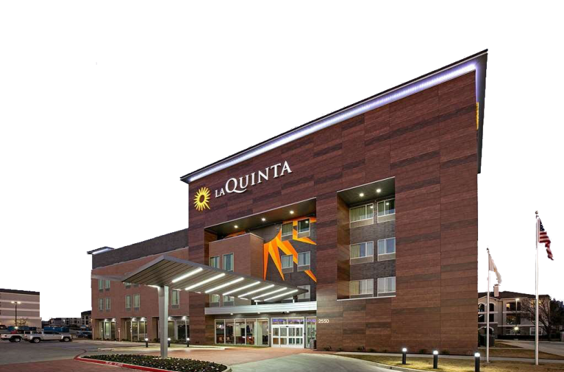 This image shows the exterior of a La Quinta hotel at night with illuminated signage and an entrance canopy. An American flag is seen on the right.