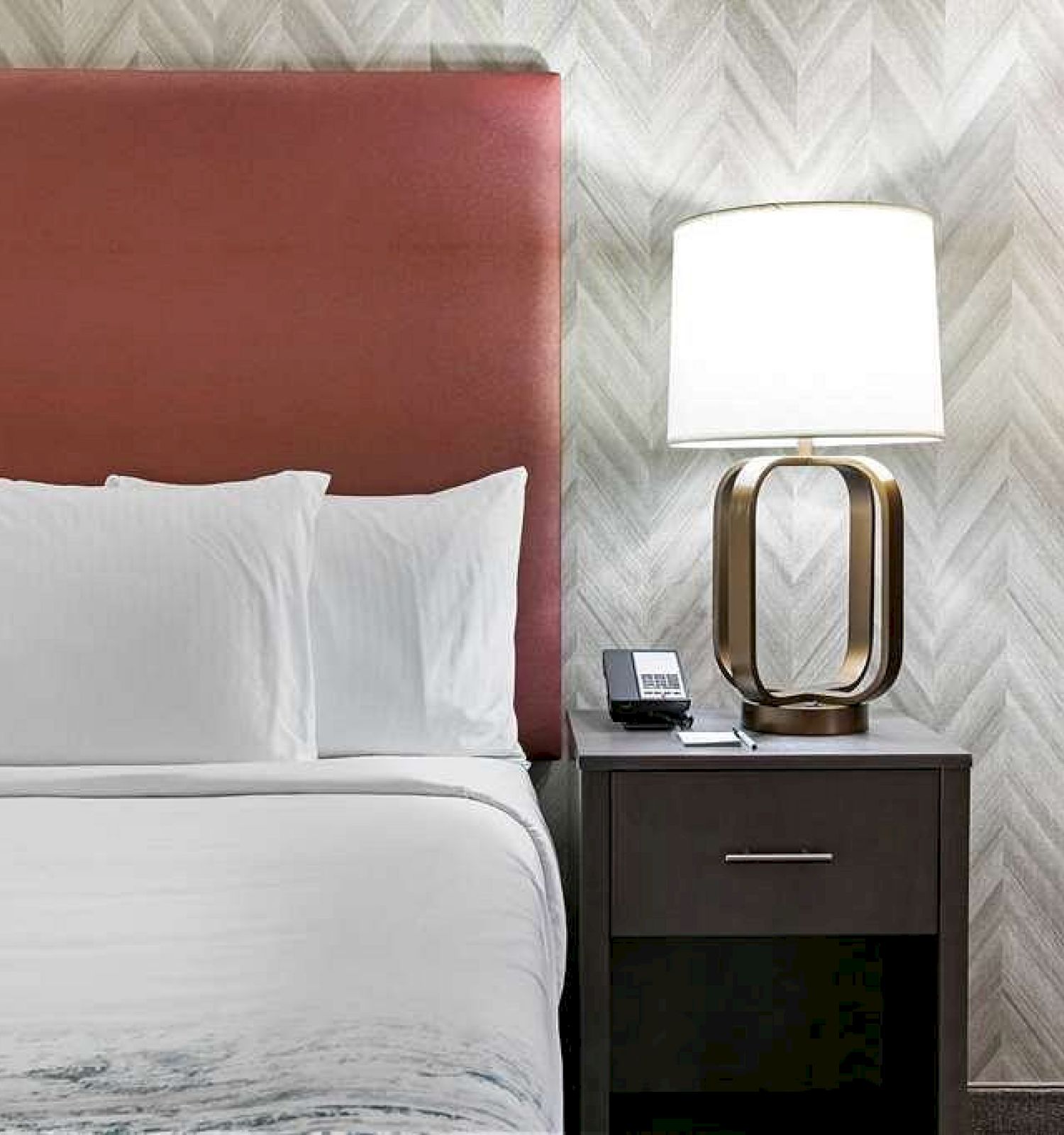 A neatly made bed with white pillows and a red headboard, next to a bedside table with a modern lamp and a phone, against a patterned wall.