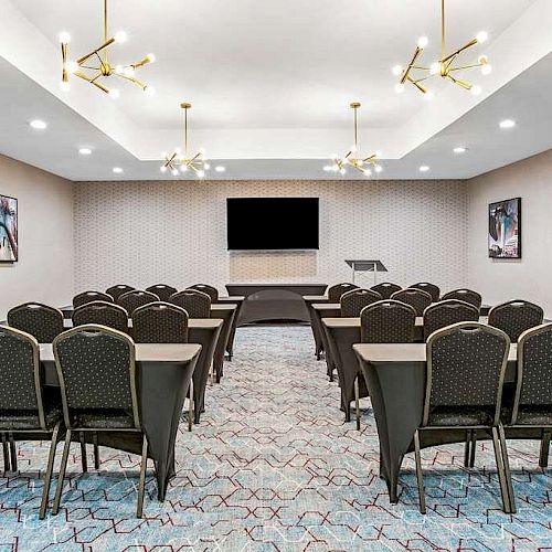 The image shows a small conference room with chairs and tables arranged in rows facing a podium and large screen. Elegant lighting decorates the ceiling.