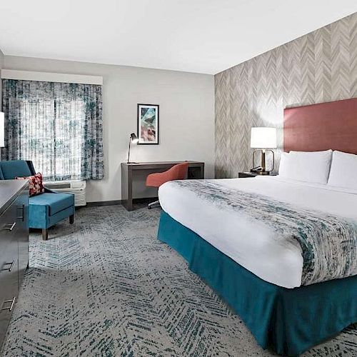 A modern hotel room with a king-size bed, a TV, an armchair, a desk, and lamps on both sides of the bed; patterned wallpaper and curtains.