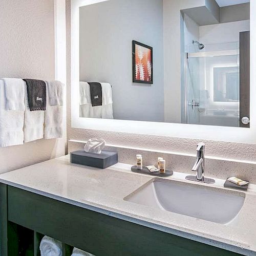 A well-lit bathroom with a large mirror, sink, faucet, soap, lotion, and tissue box on the counter, with towels hanging on the wall.