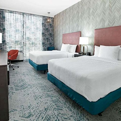 This image shows a modern hotel room with two double beds, red headboards, a TV, a desk with a chair, and large windows with curtains.