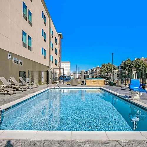 This image shows an outdoor swimming pool next to a building, with lounge chairs on one side and pool stairs on the other, under a clear blue sky.