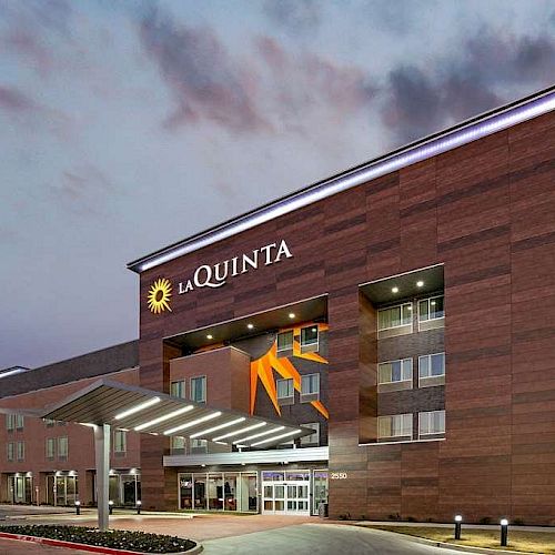 The image shows a La Quinta hotel building with a modern design at dusk, illuminated by lights, and an American flag visible on the right side.