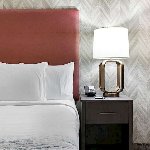 A neatly made bed with white linens, a red headboard, and a nightstand holding a modern lamp and a phone against patterned wallpaper.