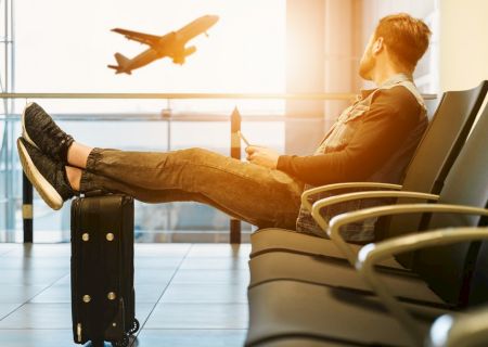 A person is sitting in an airport terminal with feet on a suitcase, watching a plane take off through the window.