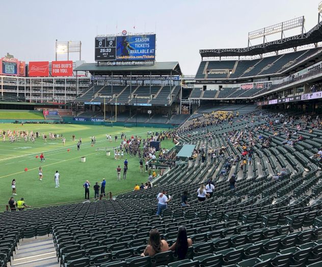 The image shows a sports stadium with a field, some spectators in the stands, and people on the field. The scoreboard has visible text.