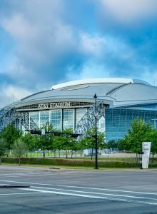 The image shows a large, modern enclosed stadium with a curved roof, surrounded by trees and an empty street in the foreground.