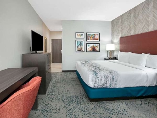 The image shows a modern hotel room with a large bed, TV, desk with a chair, bedside tables, and artwork on the walls.