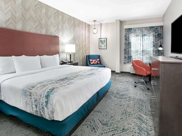 This image shows a modern hotel room with a king-size bed, a desk with a TV, an armchair, a floor lamp, and a window with curtains.