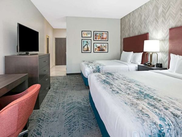 A hotel room with two beds, a TV on a dresser, a desk with a red chair, a lamp, and wall art, featuring modern decor and neutral colors.