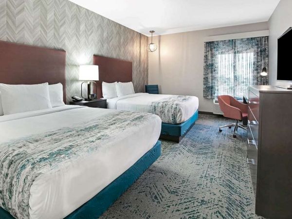 This image shows a modern hotel room with two queen beds, a TV, a desk, a chair, and a window with patterned curtains.