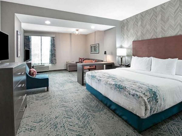A modern hotel room with a large bed, couch, TV, desk, and window. It has stylish decor with neutral colors and a patterned carpet.