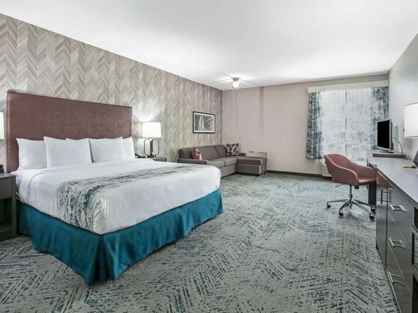This image shows a spacious hotel room with a king-sized bed, a sofa, desk with a chair, flat-screen TV, lamps, and patterned carpet flooring.