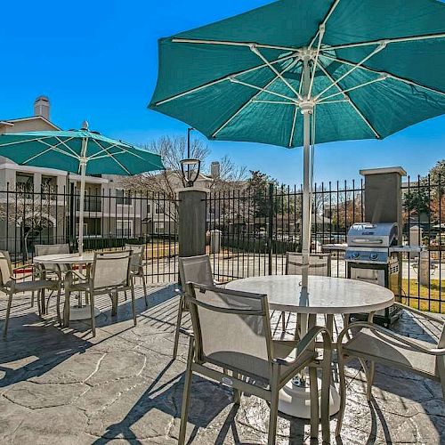 The image shows an outdoor patio area with tables, chairs, and large green umbrellas, adjacent to a fenced residential complex and a grill.