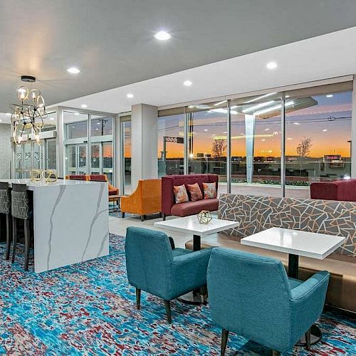 A modern lounge area with various seating options, colorful decor, large windows showing a sunset, and a chandelier.