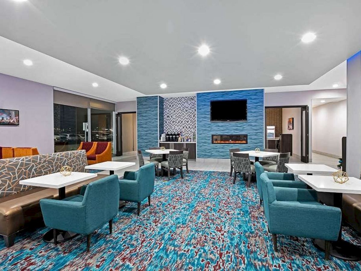 The image shows a modern hotel lounge area with blue chairs, patterned carpets, and a TV mounted on a wall; tables and cushioned benches are arranged.