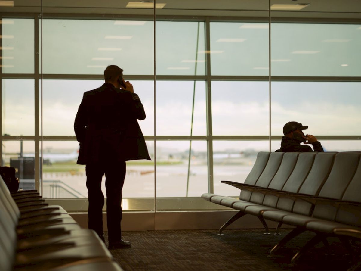 Two people are in an airport waiting area, both engaged in phone conversations, with large windows showing the runway outside.