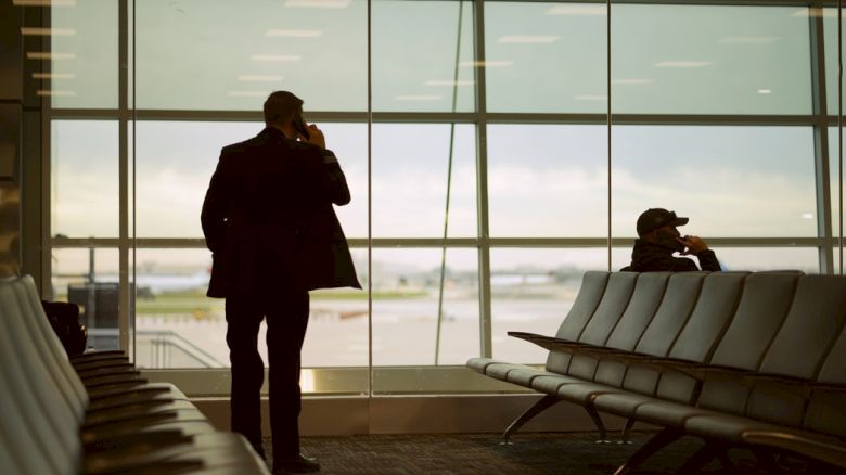 Two people are in an airport waiting area, both engaged in phone conversations, with large windows showing the runway outside.