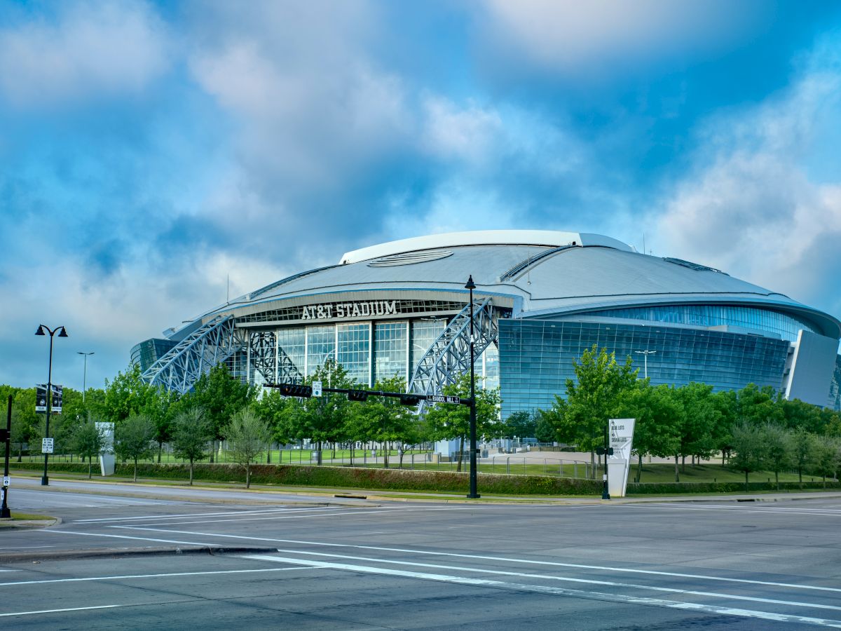 The image shows a large, modern stadium with a curved roof, surrounded by greenery and streets, with a clear, blue sky in the background.