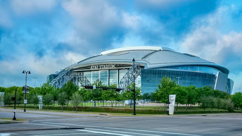 The image shows a large, modern stadium with a curved roof, surrounded by greenery and streets, with a clear, blue sky in the background.