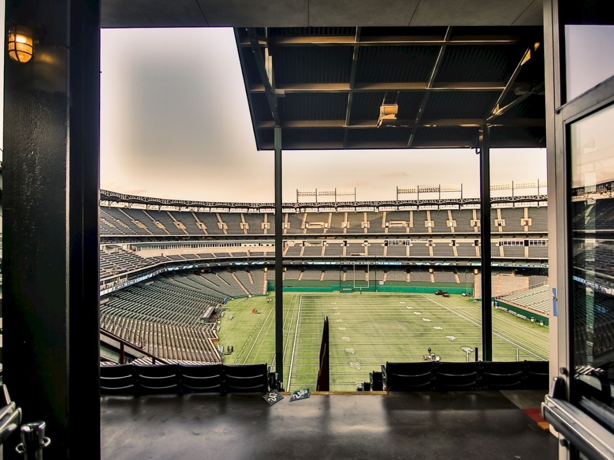 A view of a large, empty stadium is seen from a shaded interior area, revealing seating sections and a well-maintained field.