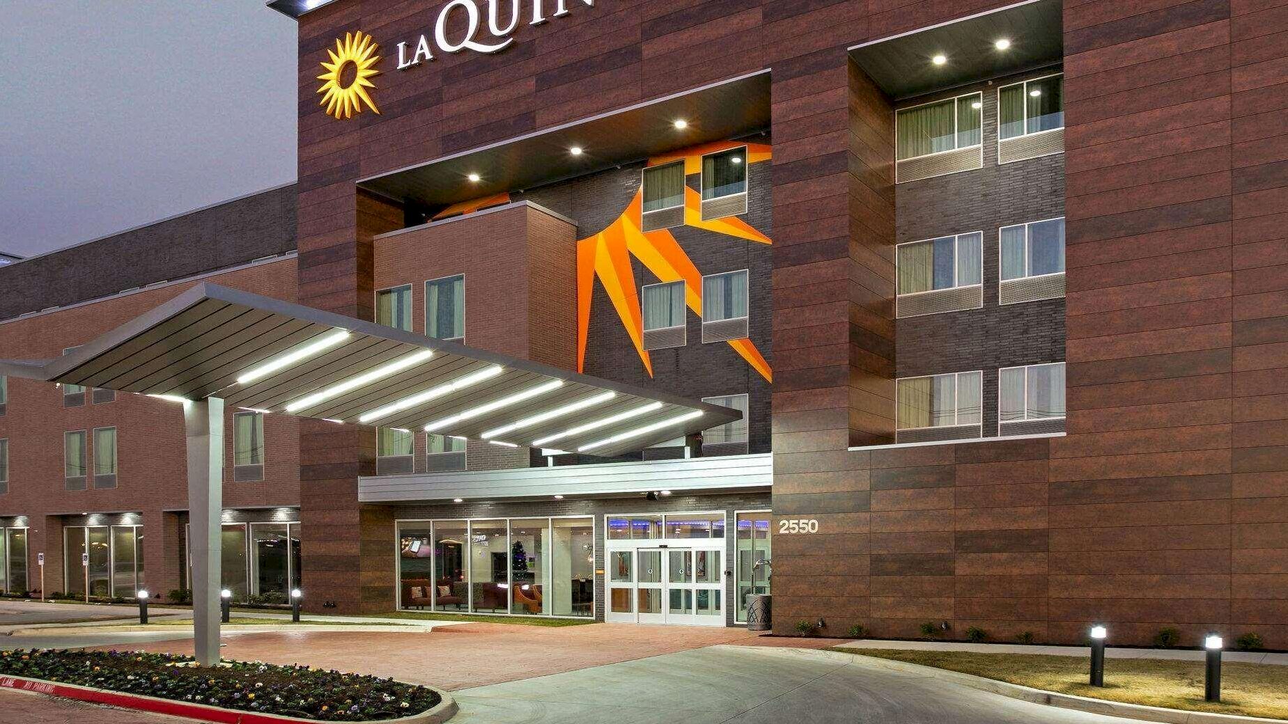 This image features the exterior of a modern La Quinta hotel at dusk, with illuminated signs, a covered entrance, and an American flag flying nearby.